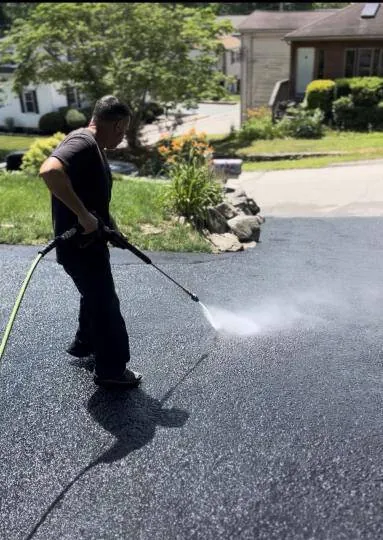 A man in a black T-shirt pressure washing a black asphalt driveway, spraying water across the surface with a wand
