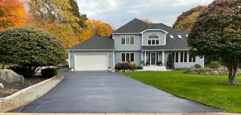Freshly sealcoated black asphalt driveway in front of a gray two-story house with fall trees